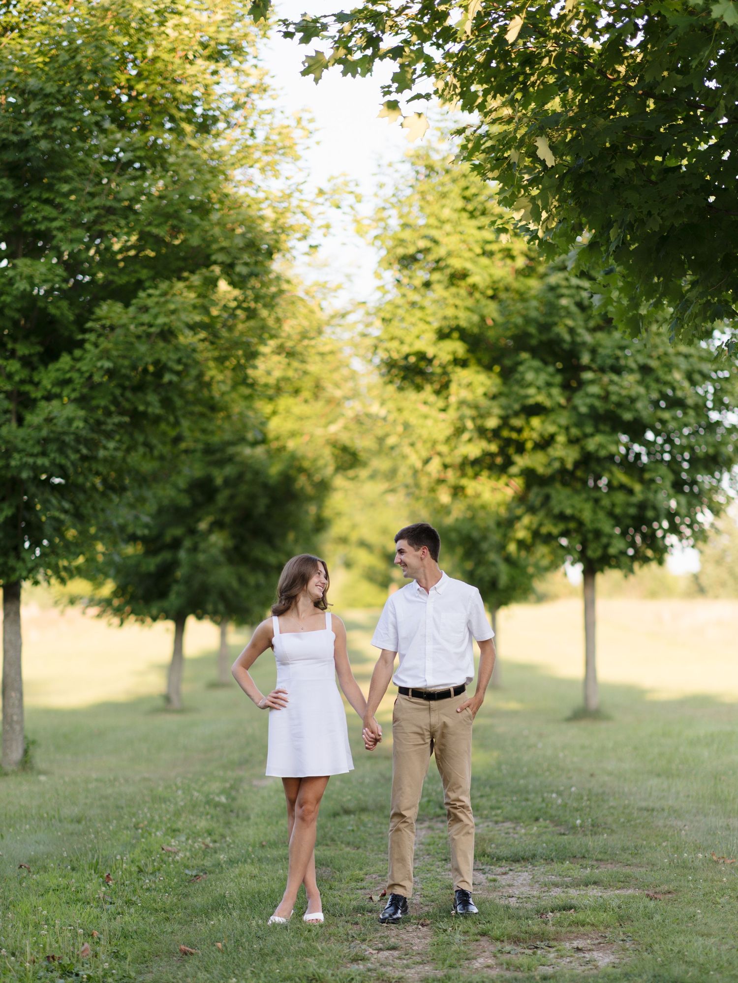 Couple holding hands smiling at each other on a tree-lined path in a sunny park.