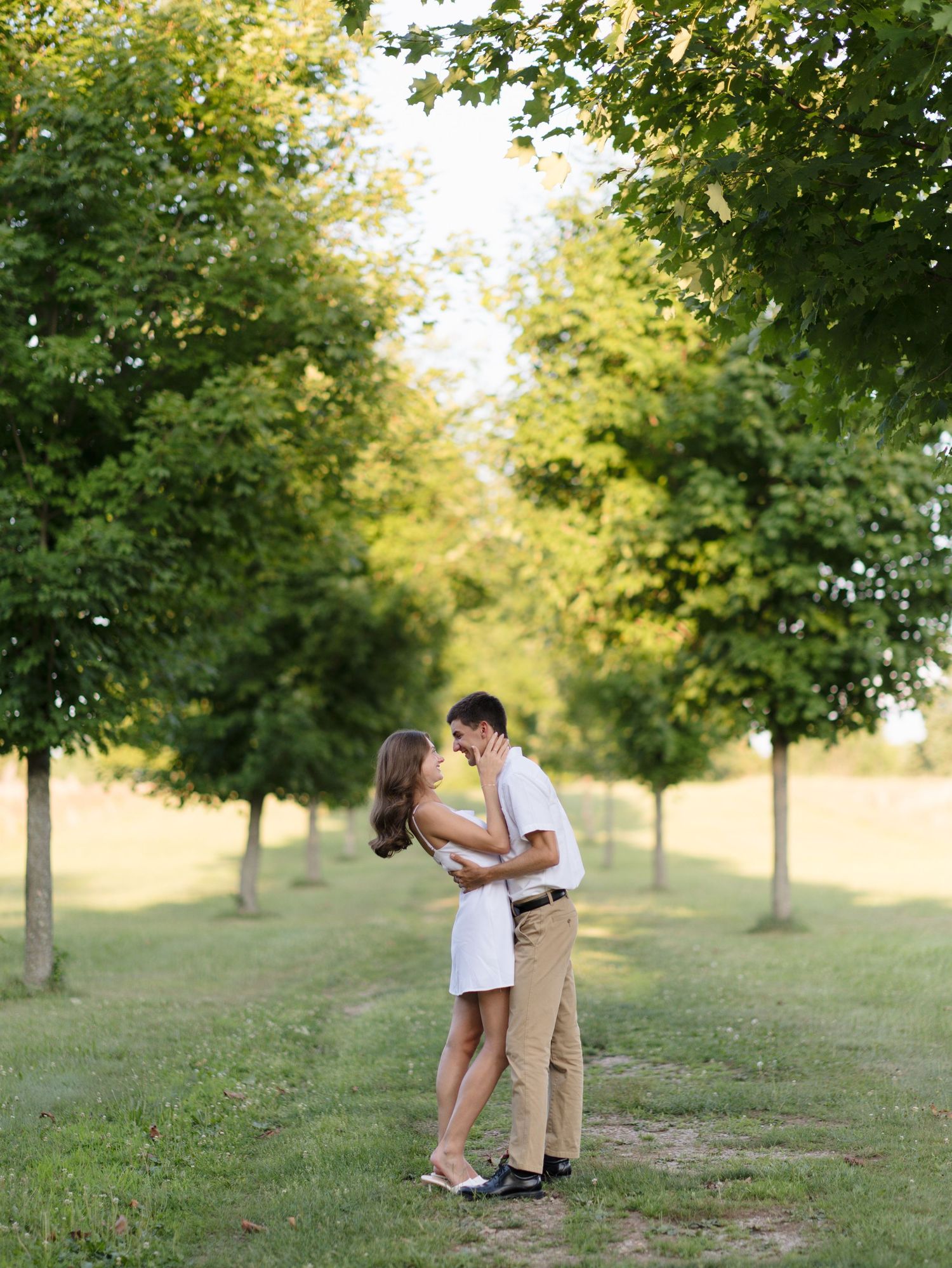 Romantic couple embracing and almost kissing between rows of green trees outdoors.
