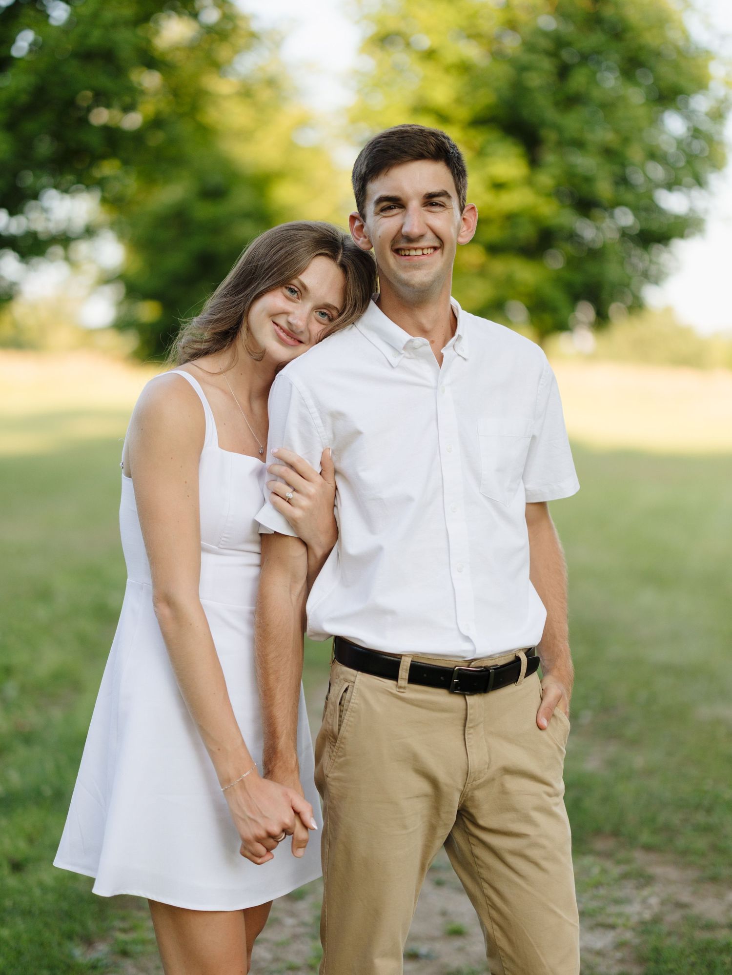 A smiling couple in white outfits poses together in a sunny park, holding hands among lush green trees.