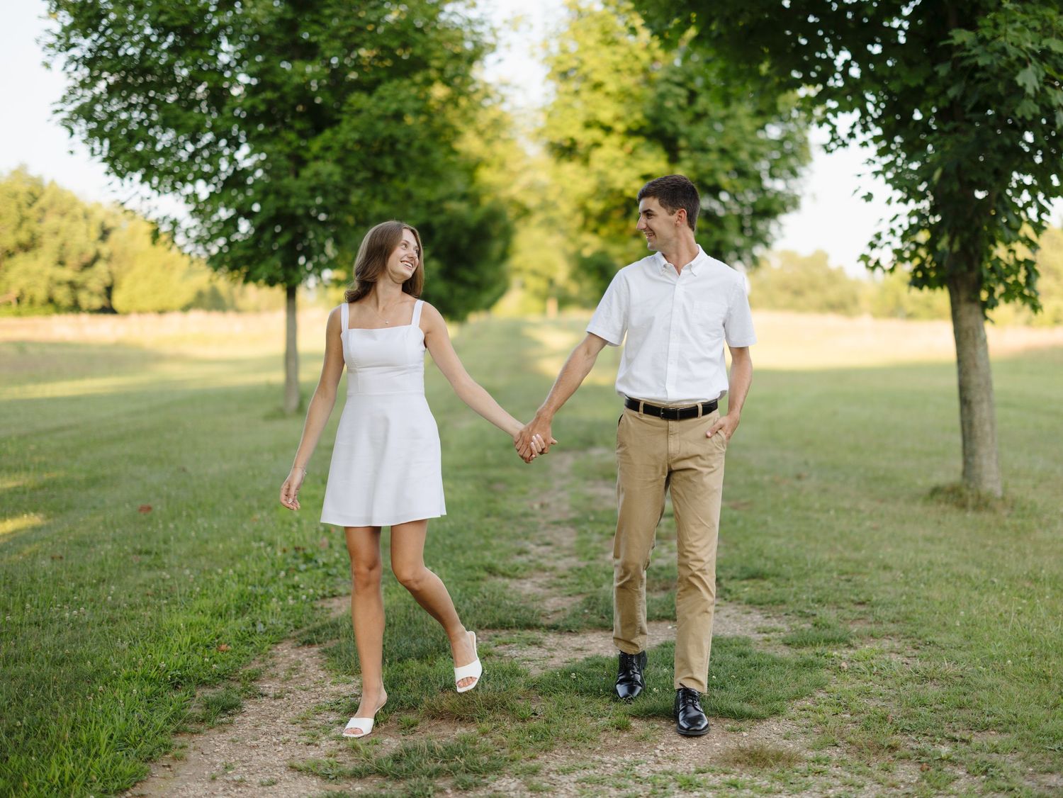 A couple holds hands while walking along a tree-lined path in a sunny green park.