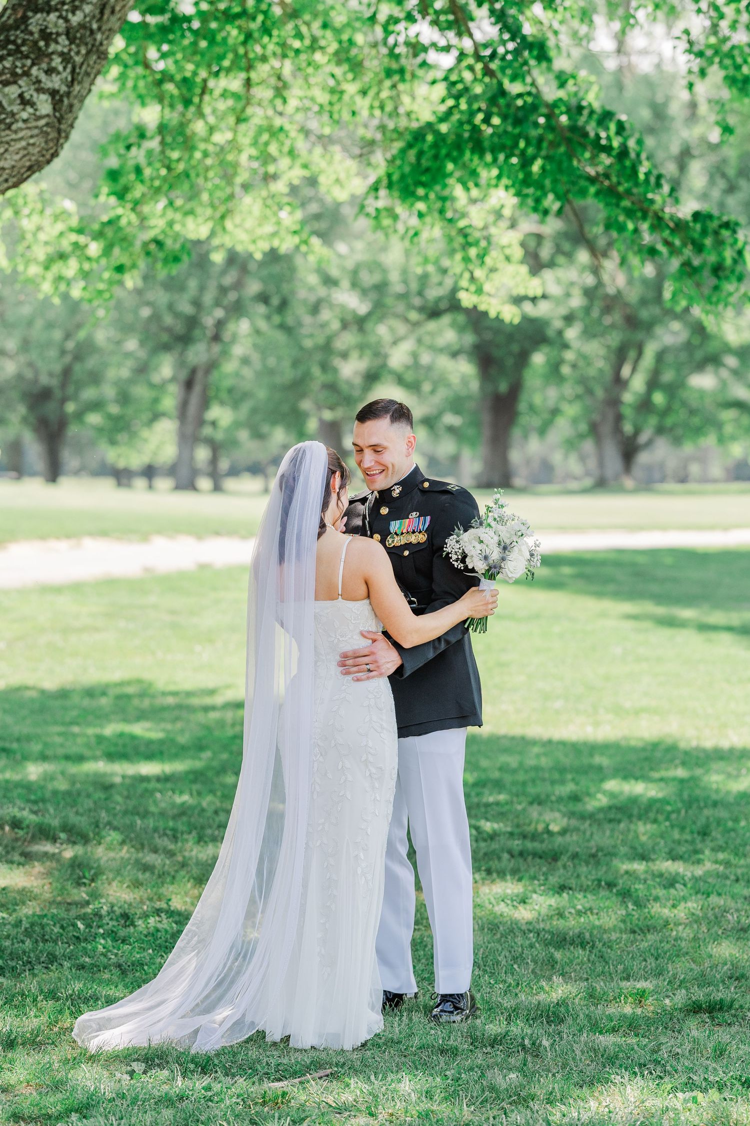 Military groom in dress uniform embraces bride in white gown and veil outdoors under green trees on wedding day.