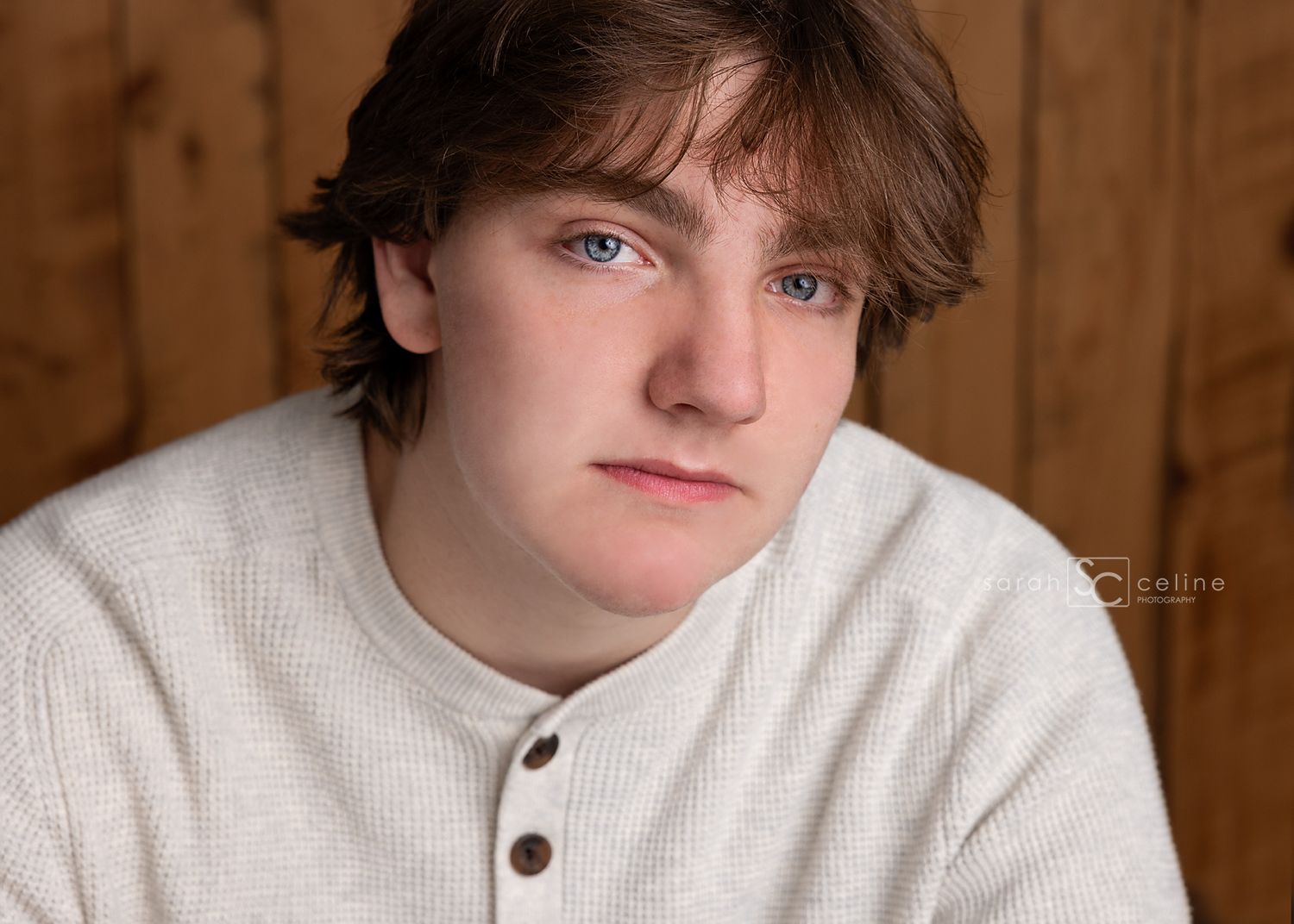 Teen boy with blue eyes and brown hair poses for senior portrait in cream henley sweater against wood background.