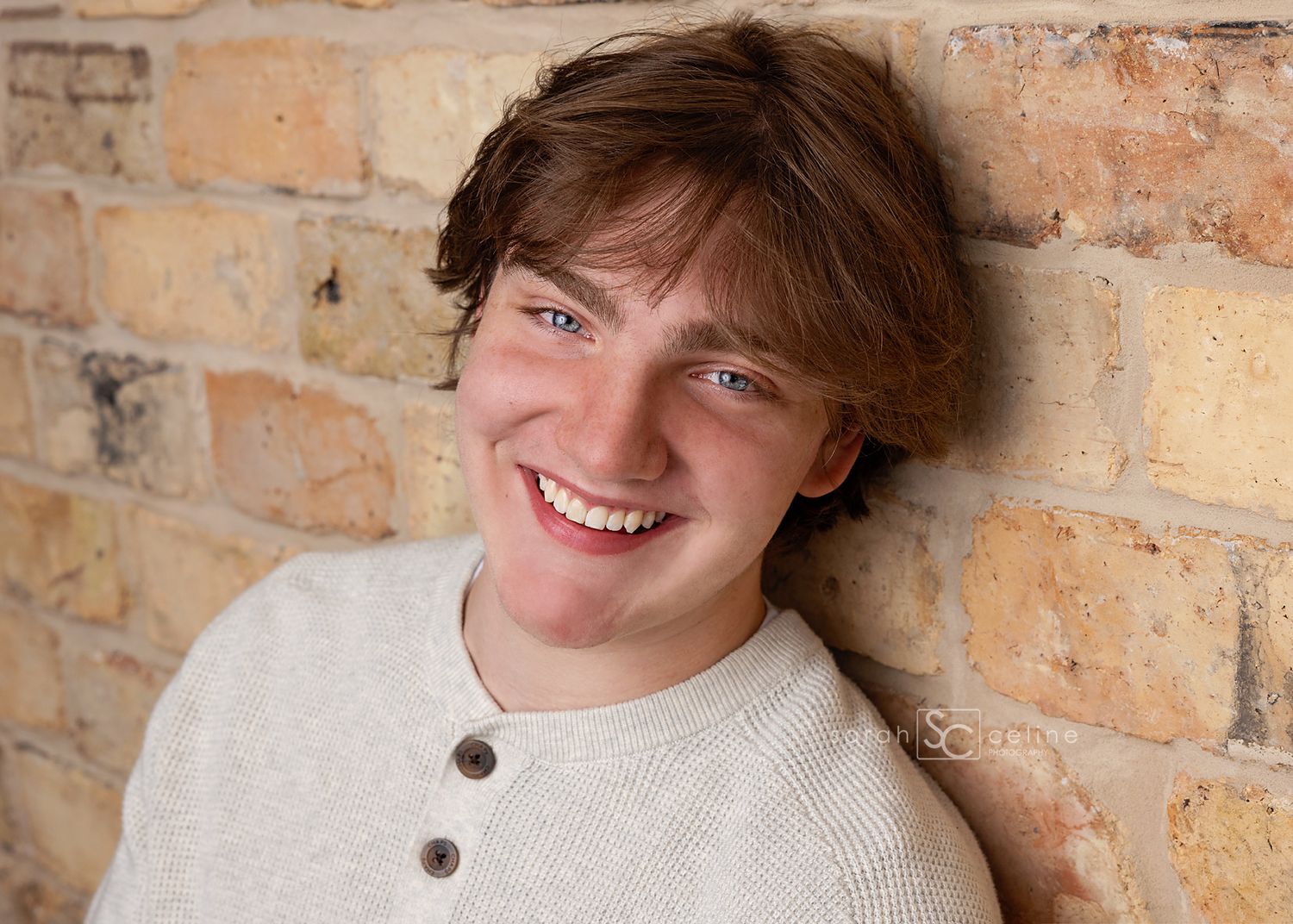 Smiling teenage boy with brown hair and blue eyes leaning against a rustic brick wall in a cream sweater.