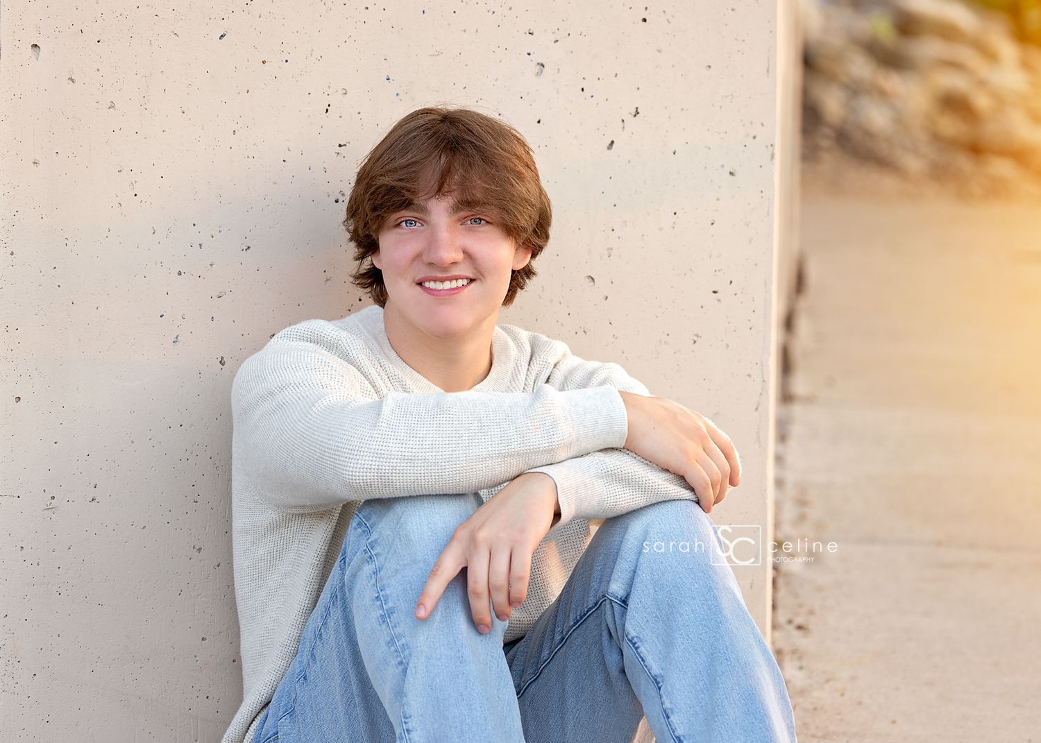 A smiling teenage boy in a white sweater and jeans sits relaxed against a concrete wall in golden hour light.