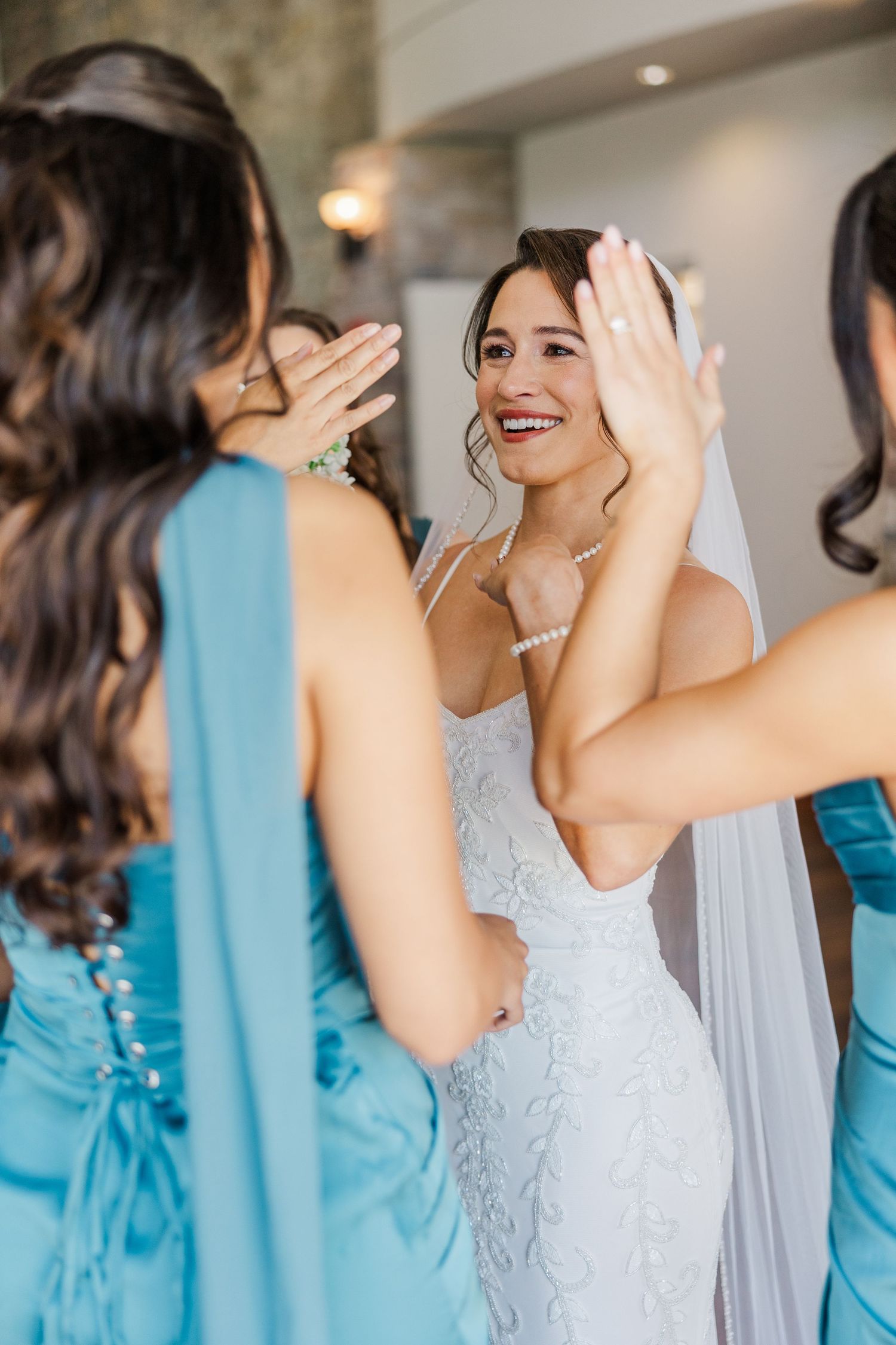Bride in white lace gown and veil shares joyful moment with bridesmaids in blue dresses before wedding ceremony.