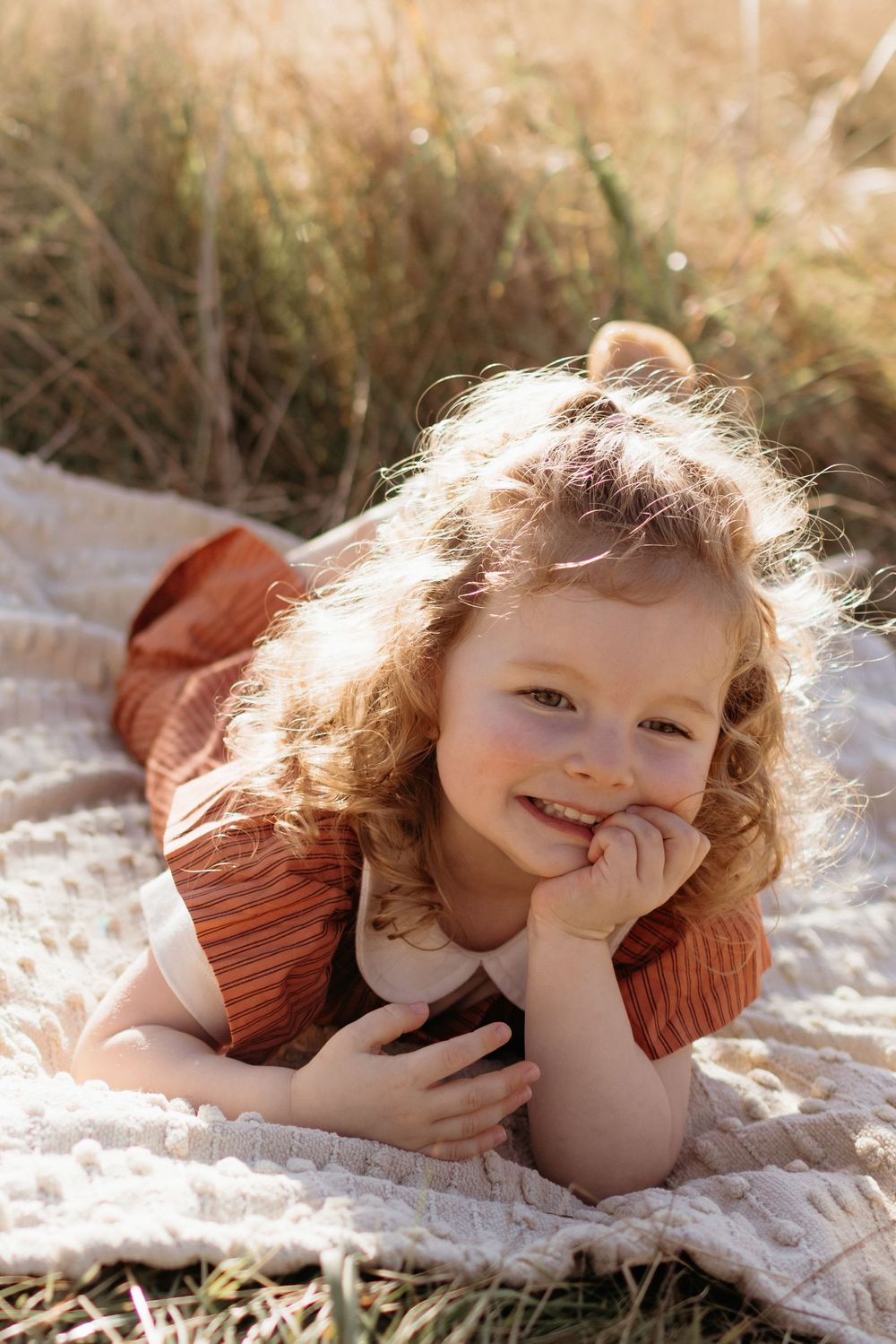 Curly-haired little girl in a rust striped dress lying on a white blanket outdoors, smiling in golden sunlight.