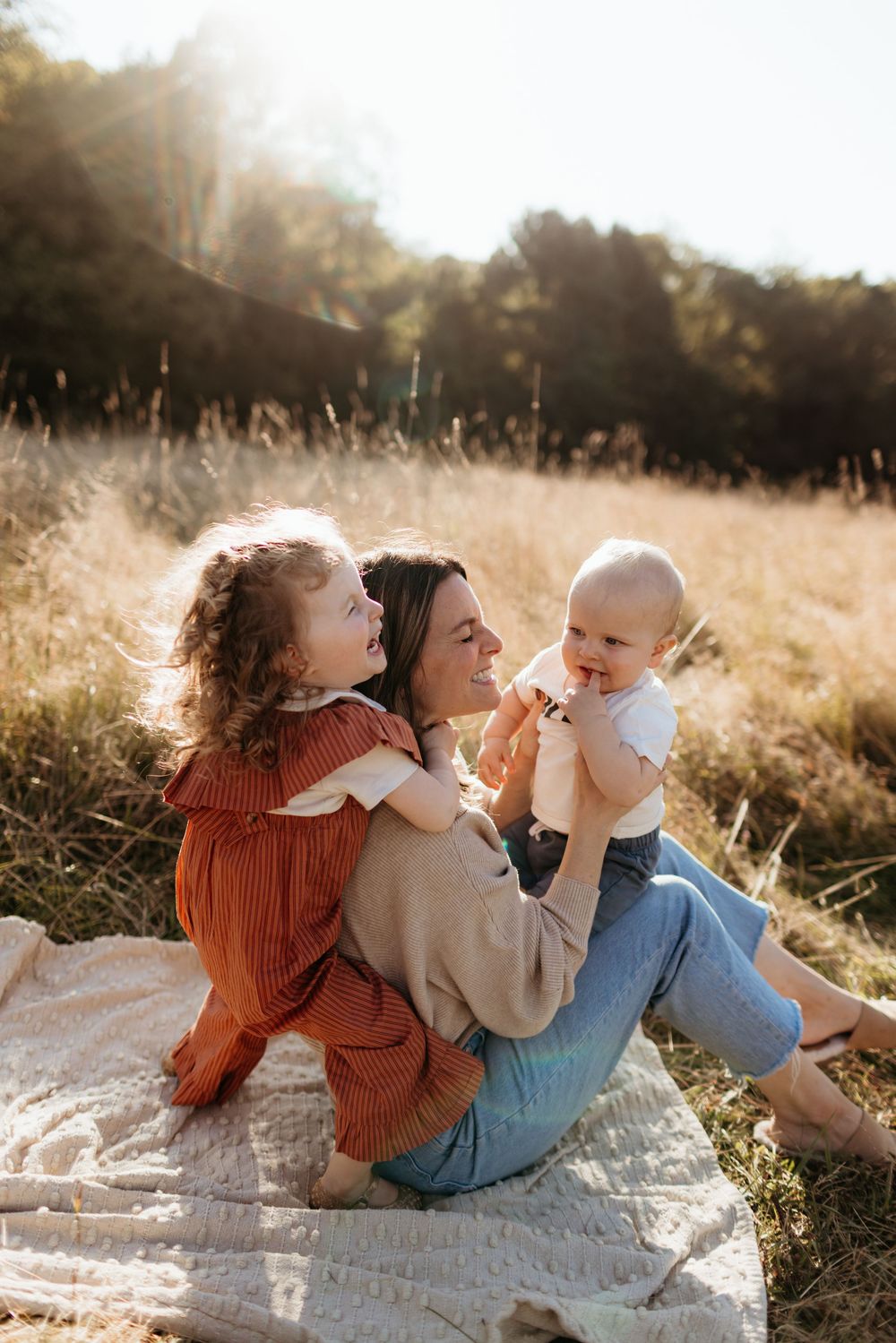 A mother sits on a blanket in a sunny golden field, smiling while holding her baby and toddler in an orange dress.
