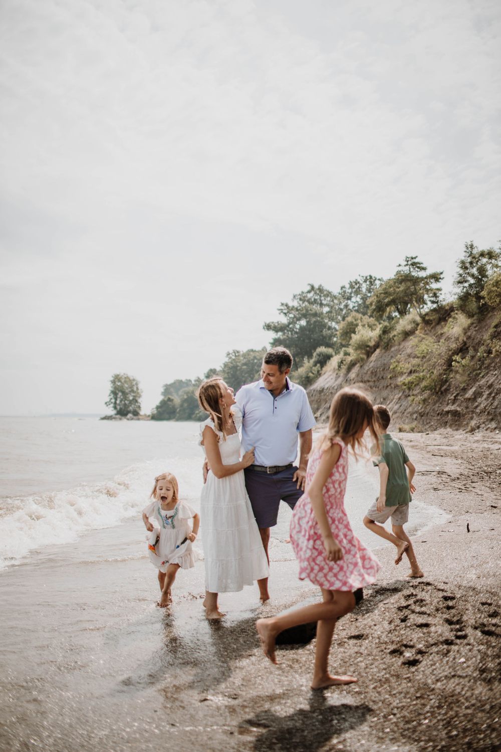 A happy family of five walks along the shoreline, splashing in the waves on a cloudy day.