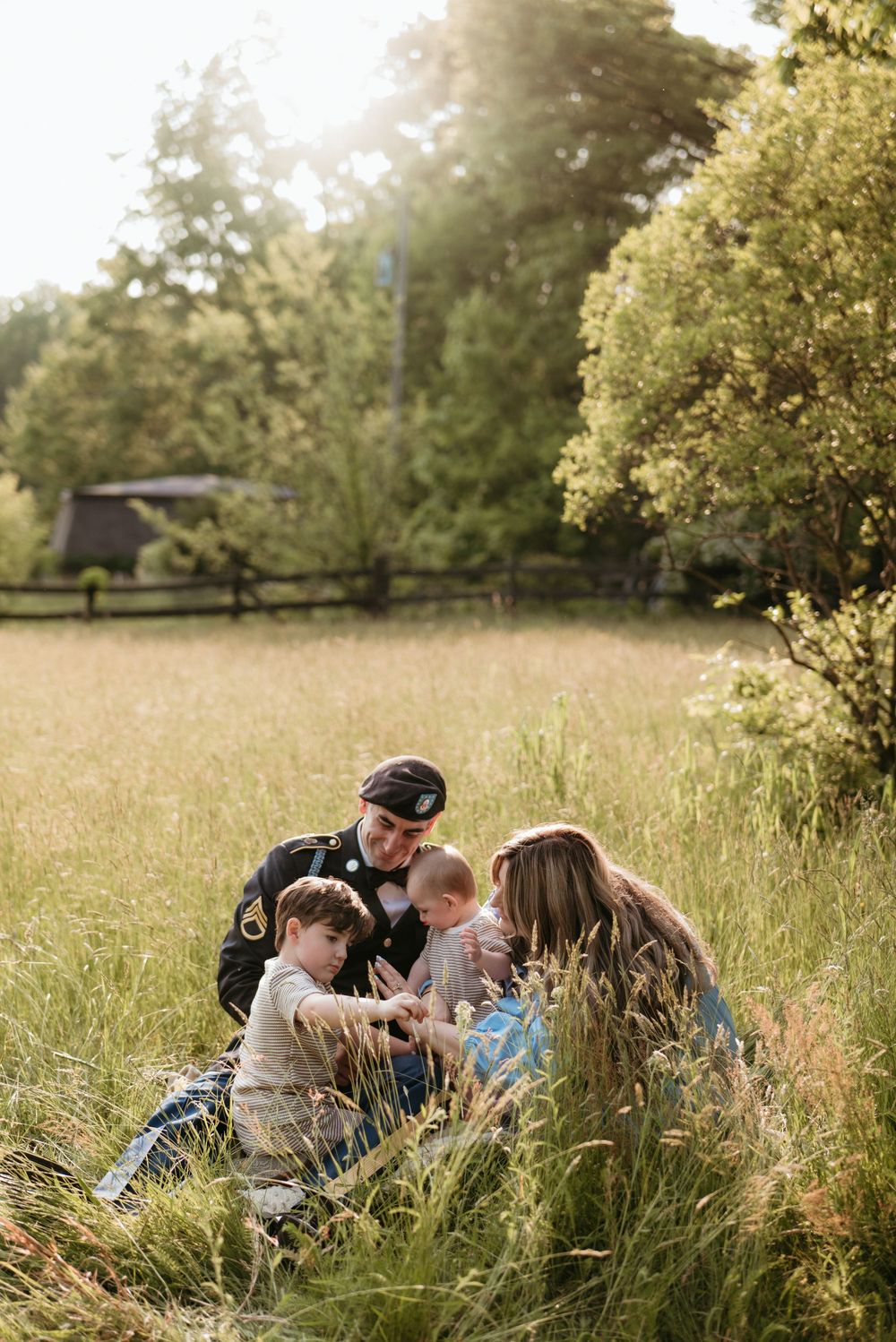 Military family sitting together in a sunlit field, sharing a tender moment with young children outdoors.