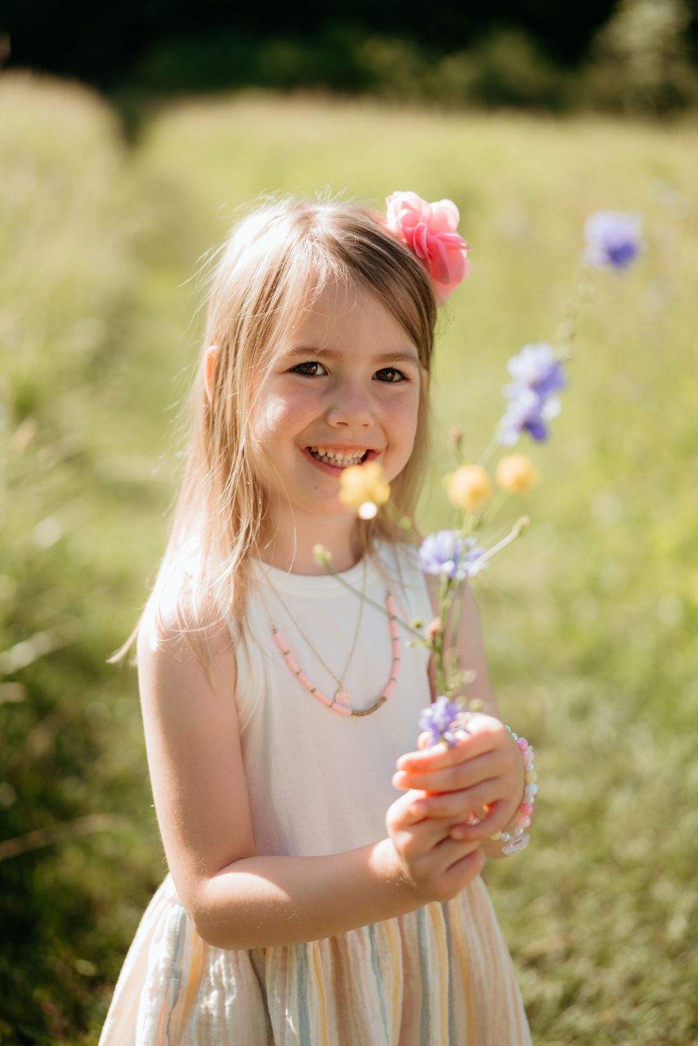 Smiling little girl in striped dress holds wildflowers with pink flower in her hair.