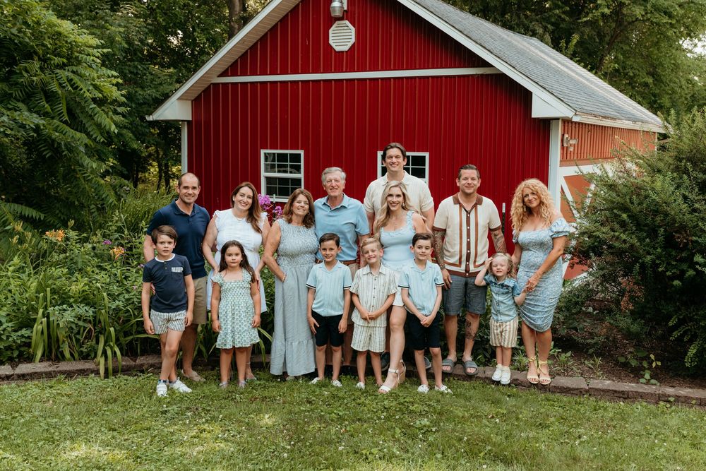 Large family group photo posing outdoors in front of a red barn on a sunny day.