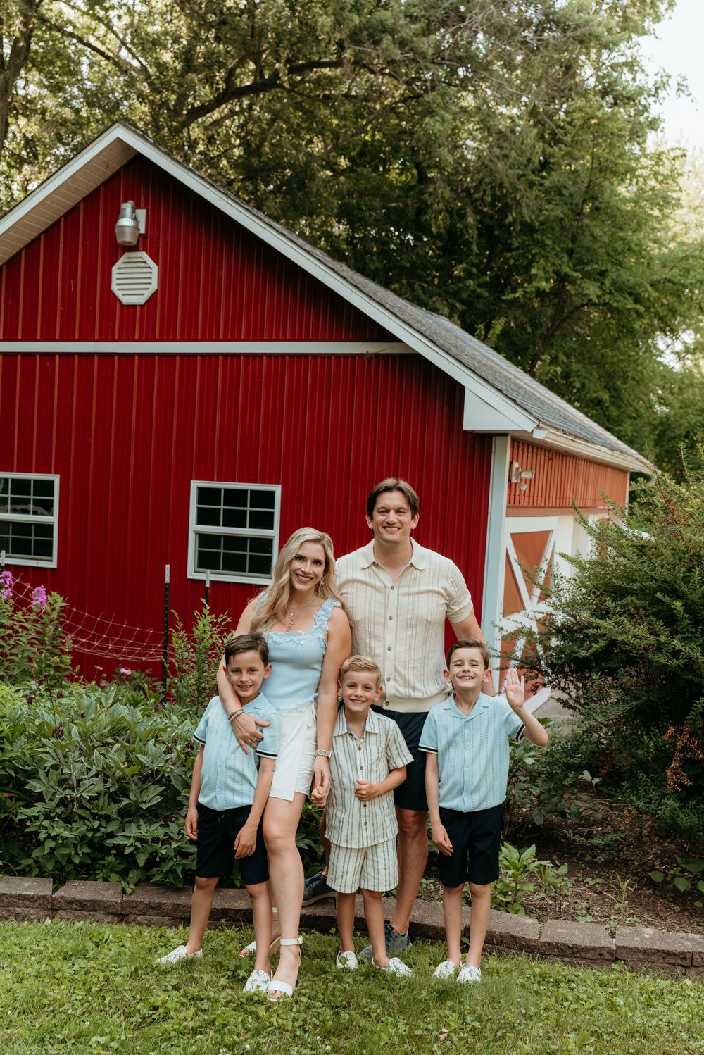 Family of five posing in front of a red barn surrounded by lush greenery, wearing coordinated blue and neutral outfits.