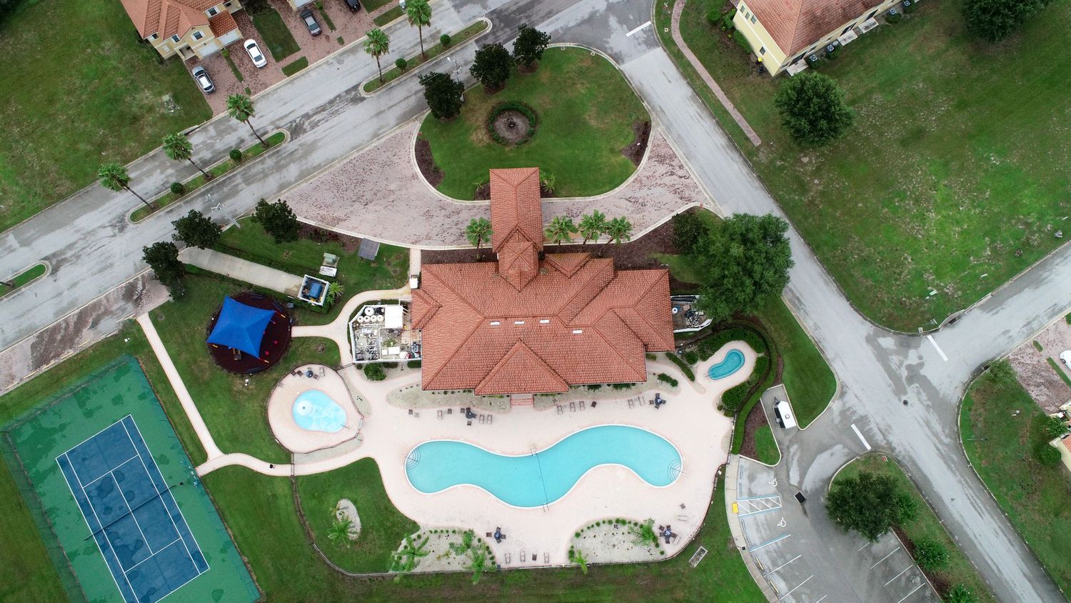 Aerial drone view of a residential home with brown shingle roof surrounded by lush green trees and a tennis court nearby.