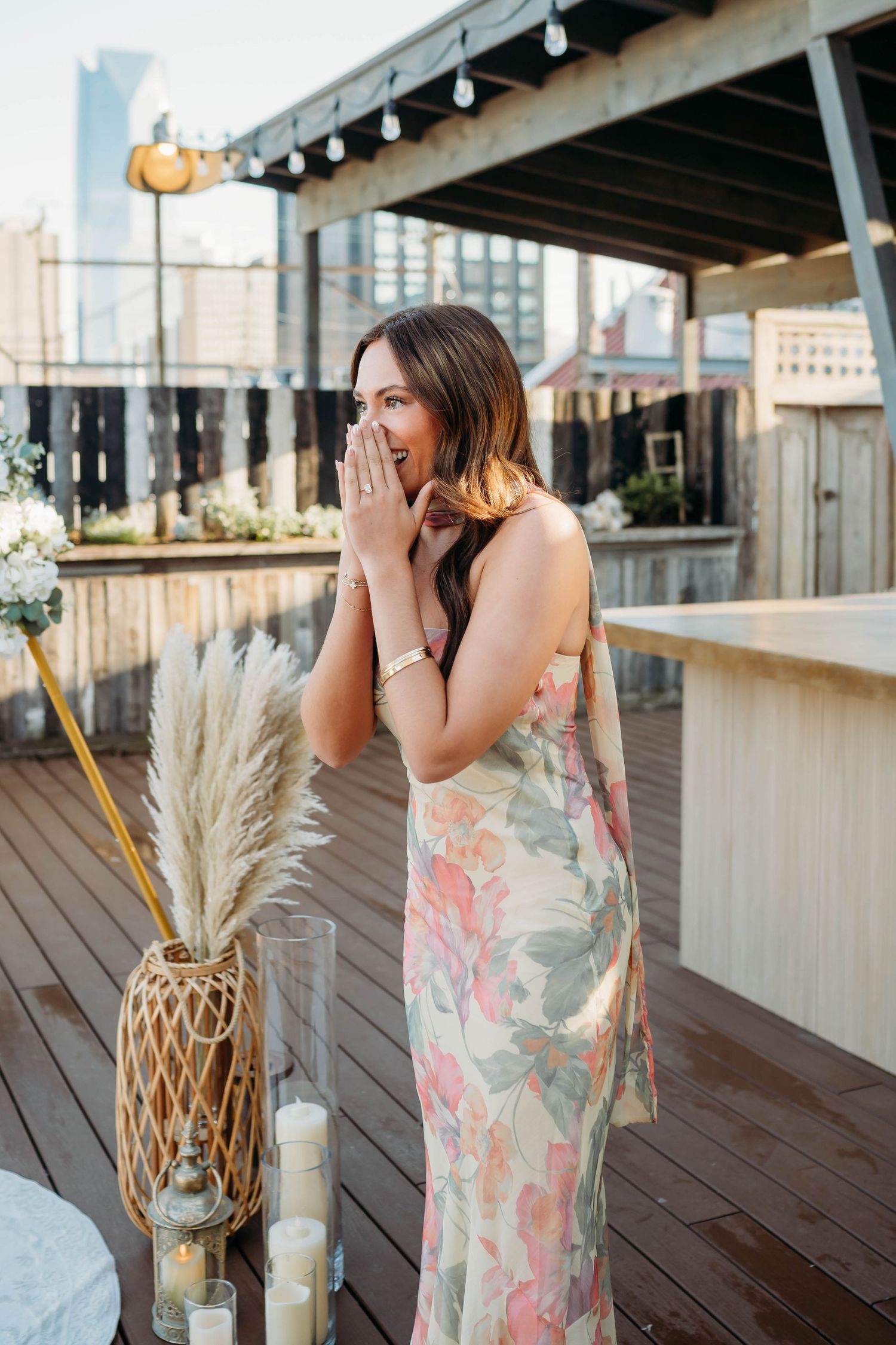 Woman in floral dress on rooftop terrace with pampas grass decor and city skyline, hands raised to mouth in surprise.