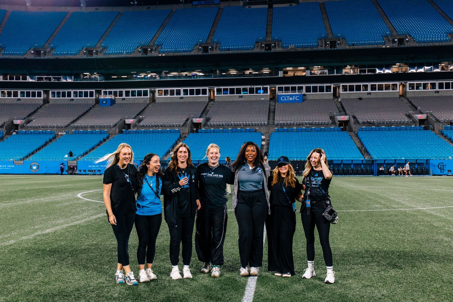Seven women pose together on a football field inside a large NFL stadium with empty blue seats.
