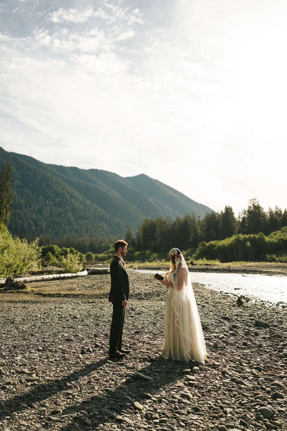 Bride and groom exchange vows on a rocky riverbank with lush green mountains in the background.