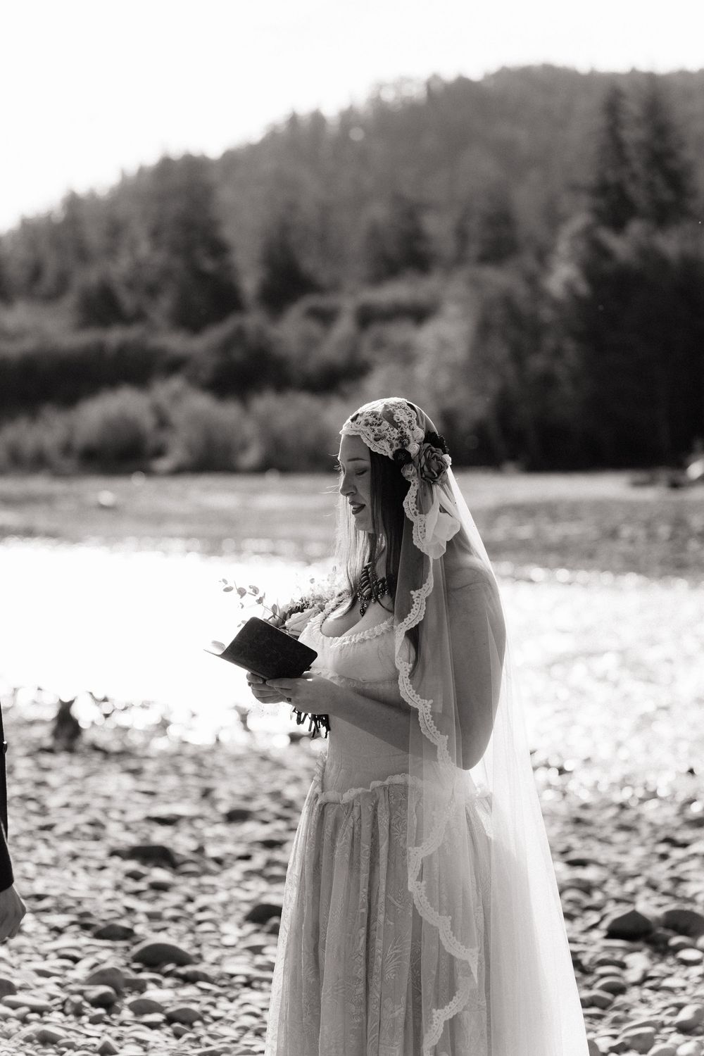 Black and white photo of a bride in a lace gown and floral crown reading vows by a river with forested hills behind her.
