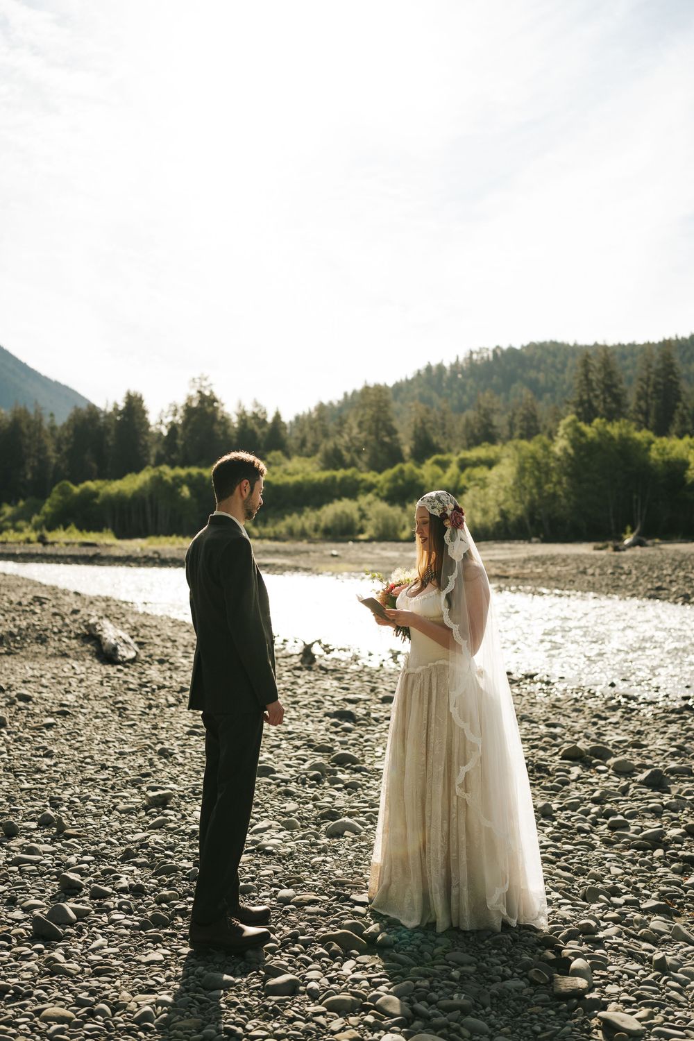 Bride reads vows to groom by a rocky riverside with mountains and forest in background during outdoor wedding ceremony.