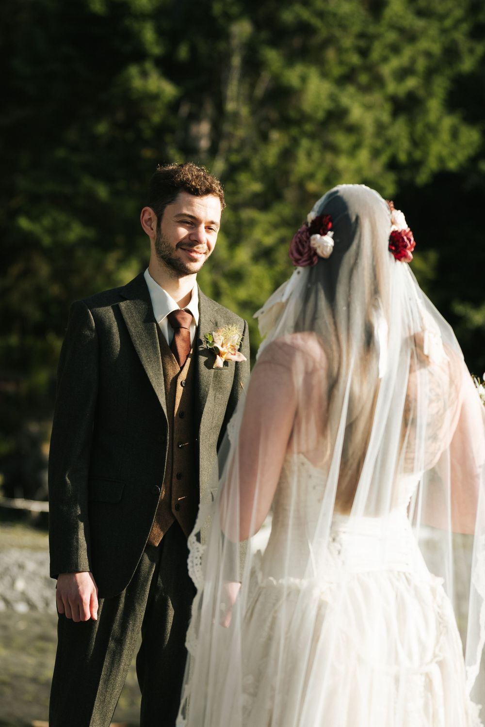 Smiling groom in gray suit with boutonniere faces bride in white lace gown and floral veil outdoors.