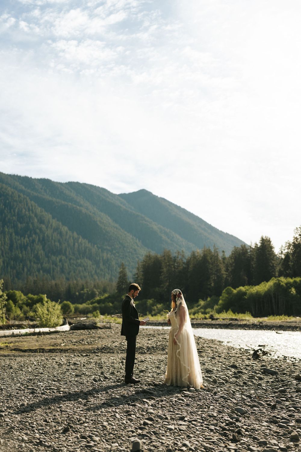 Bride and groom face each other on a rocky riverbank with lush green mountains and pine forest in the background.