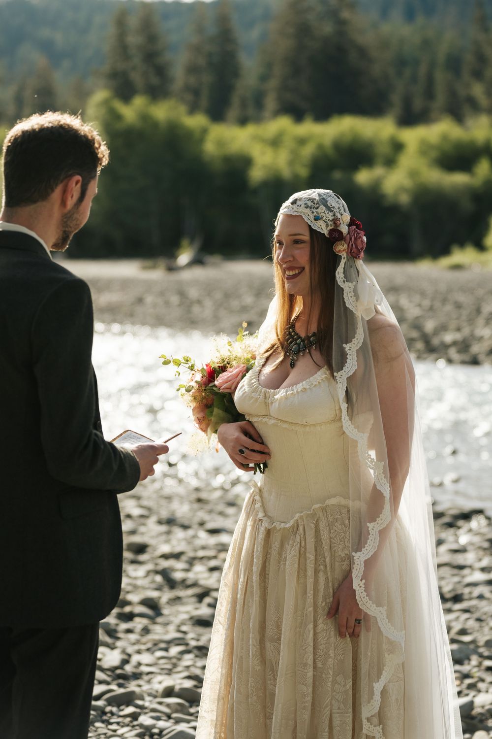 Groom reads wedding vows to smiling bride in vintage lace dress and floral veil by a mountain river.