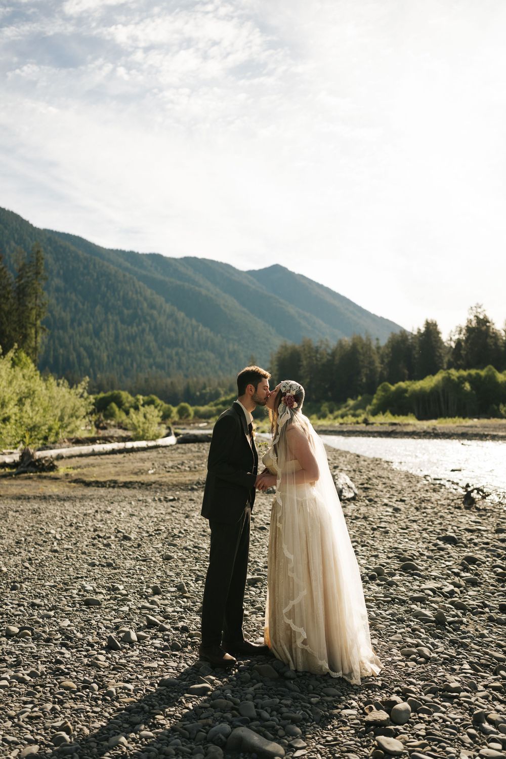 A bride and groom share a kiss on a rocky riverbank with lush green mountains in the background.