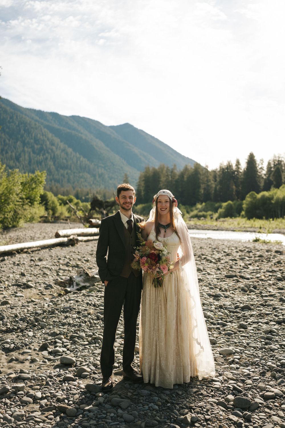 Bride and groom posing on rocky riverbank with mountain forest backdrop on their wedding day.
