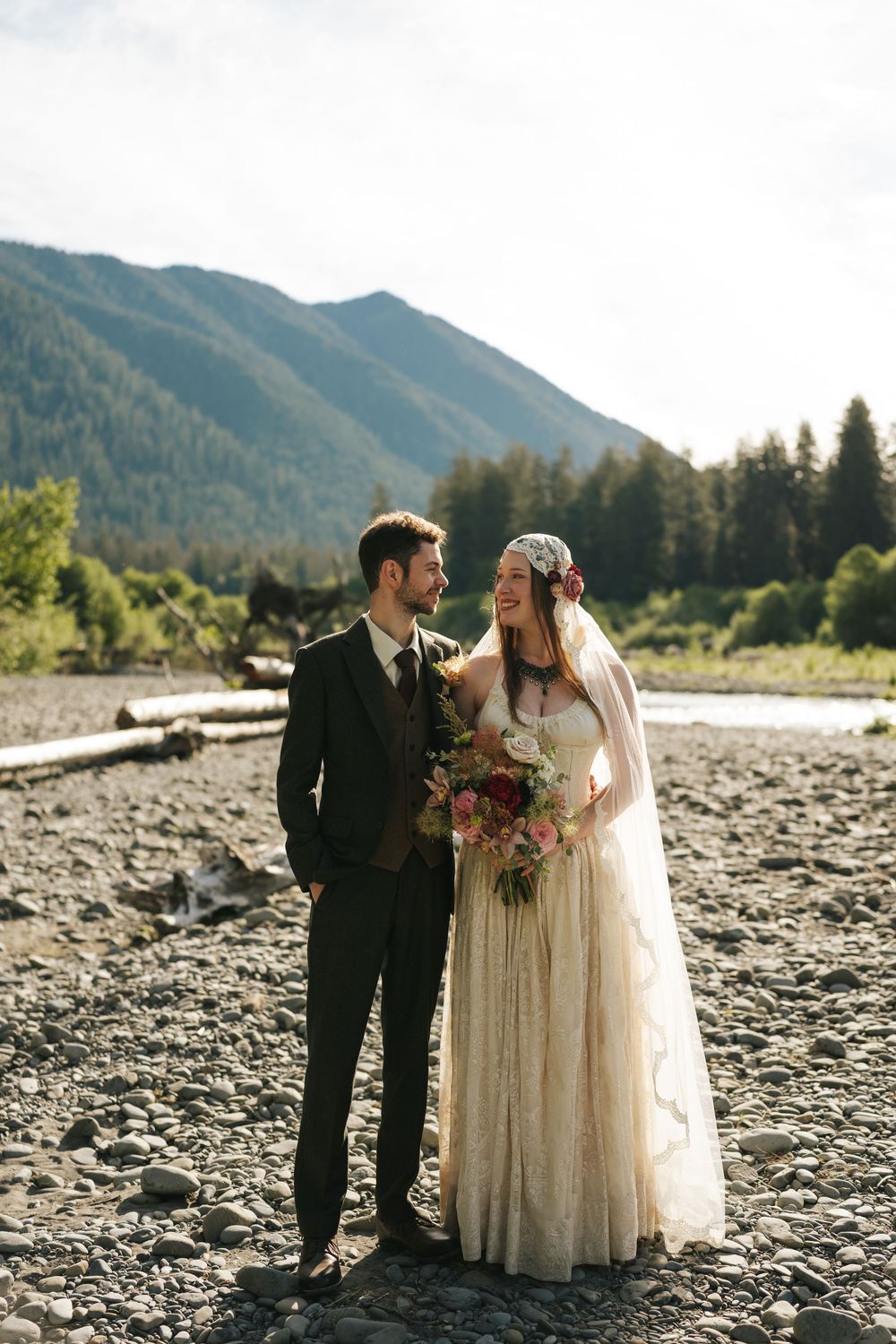 Bride and groom standing on rocky riverbank with mountain forest backdrop on their wedding day.