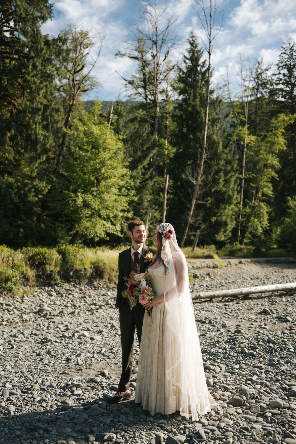 Bride holding bouquet and groom standing on rocky shore with lush green forest and mountains behind.