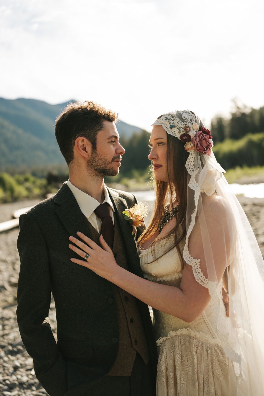 A bride in a lace veil with flowers and groom in a dark suit share an intimate moment outdoors.