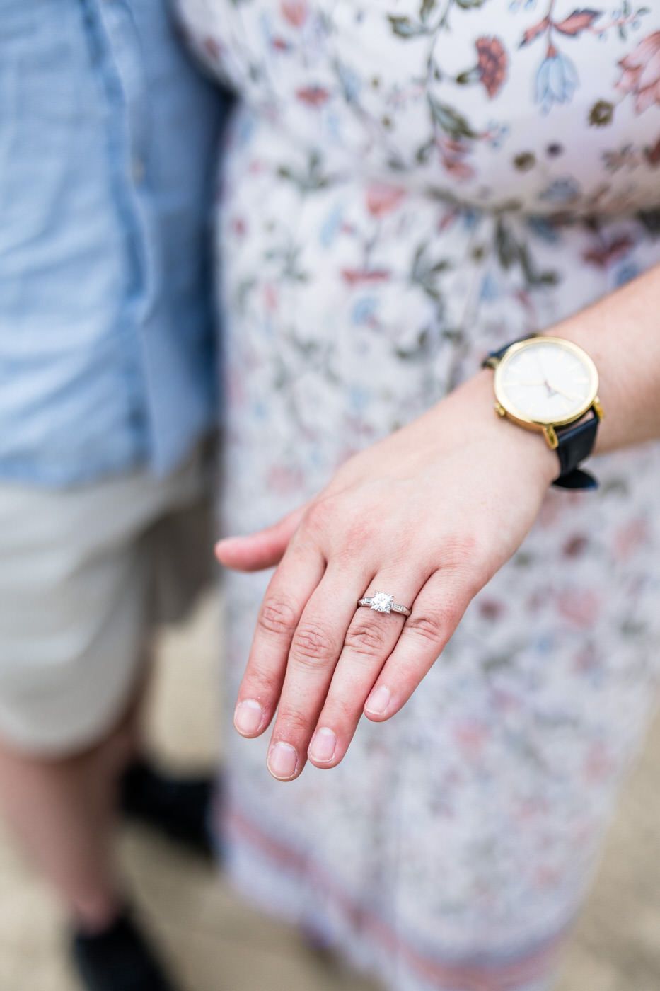 Close-up of woman's hand displaying diamond engagement ring and gold watch, with floral dress and man in blue shirt.