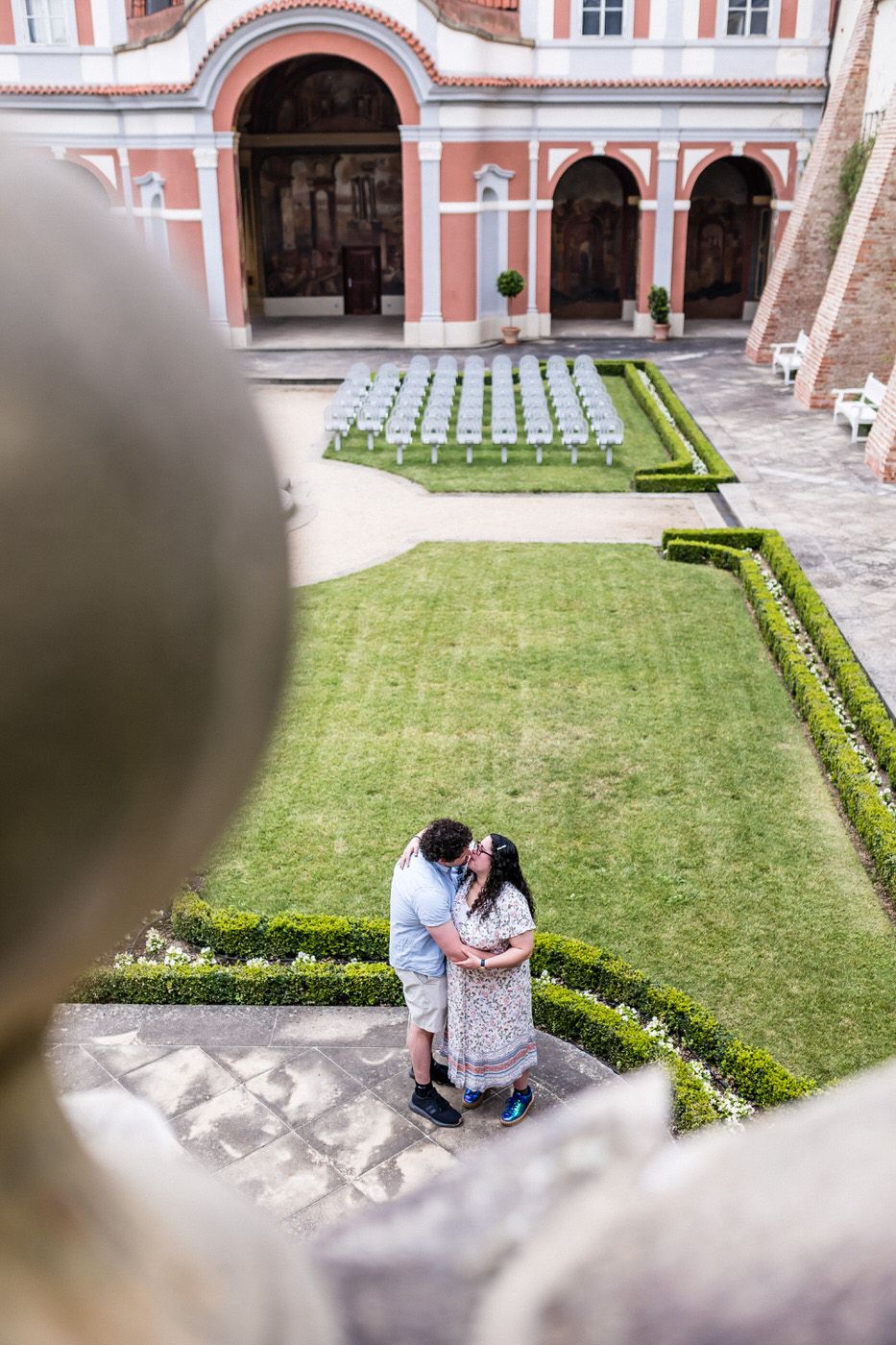 Couple sharing a romantic kiss in an elegant courtyard garden with manicured hedges and white chairs.