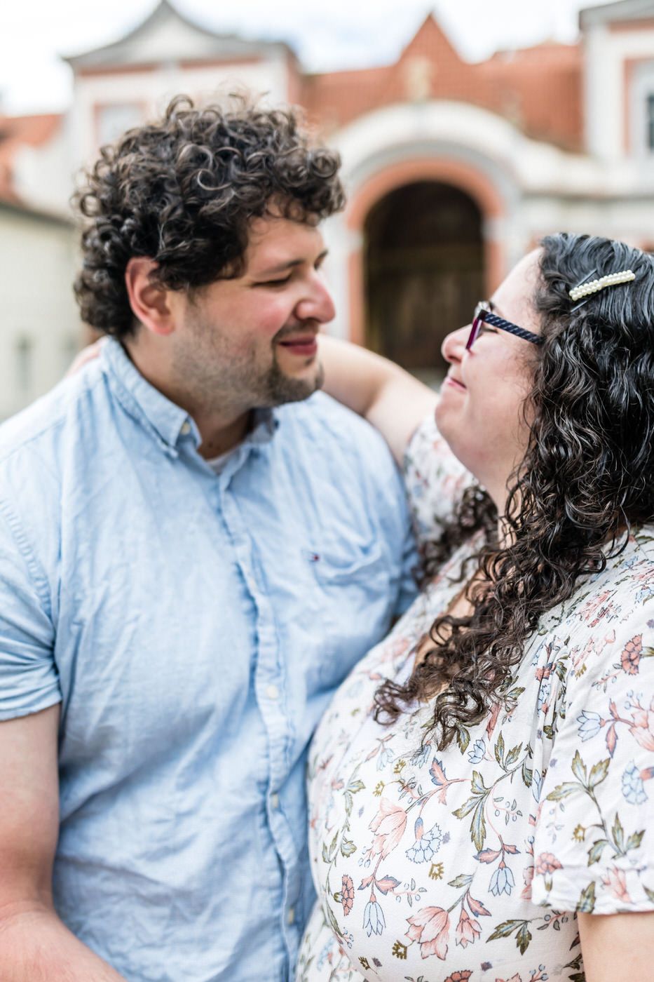 A curly-haired couple smiling at each other outdoors, woman wearing glasses and floral dress, man in light blue shirt.