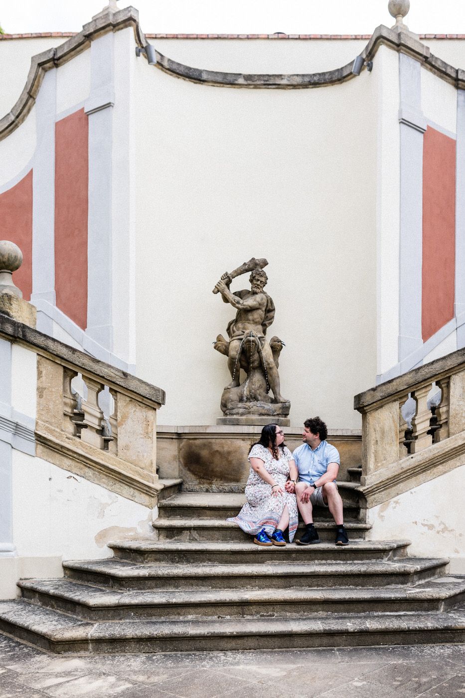 Couple sitting on ornate baroque staircase with classical sculpture, beside a red and white historic building.