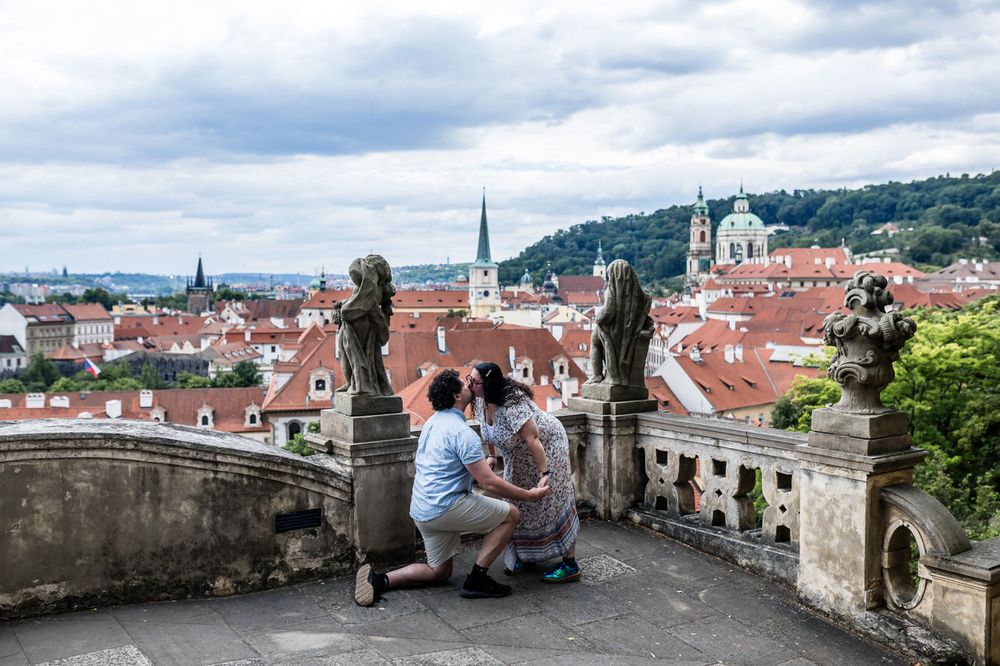 Couple sitting on a scenic terrace overlooking Prague's red rooftops and historic church spires.