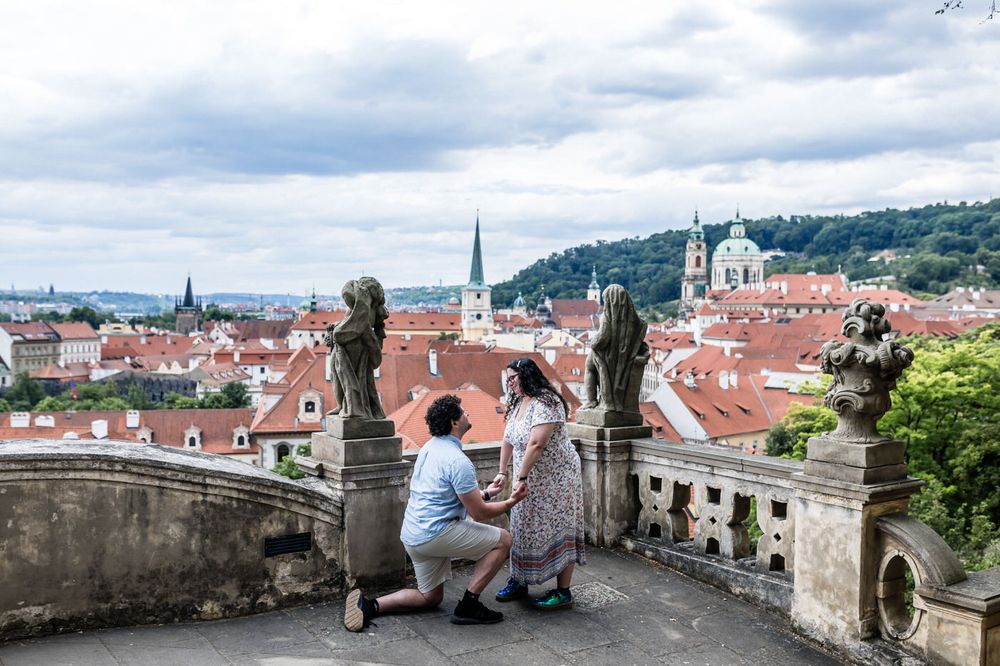Couple sharing a romantic proposal moment on a scenic Prague terrace overlooking red rooftops and historic church spires.