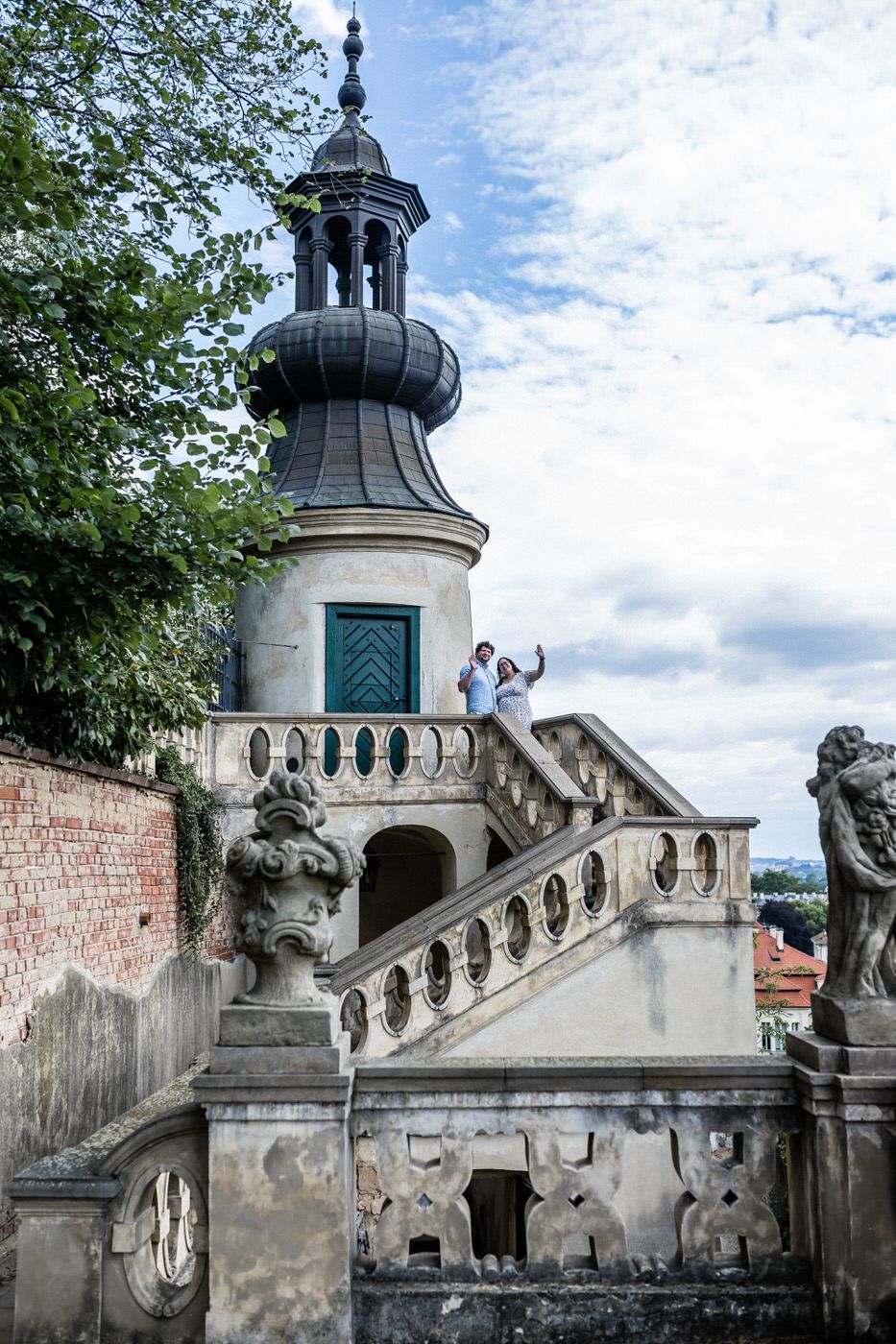 Couple posing on ornate baroque staircase by decorative tower with statues at historic European castle.