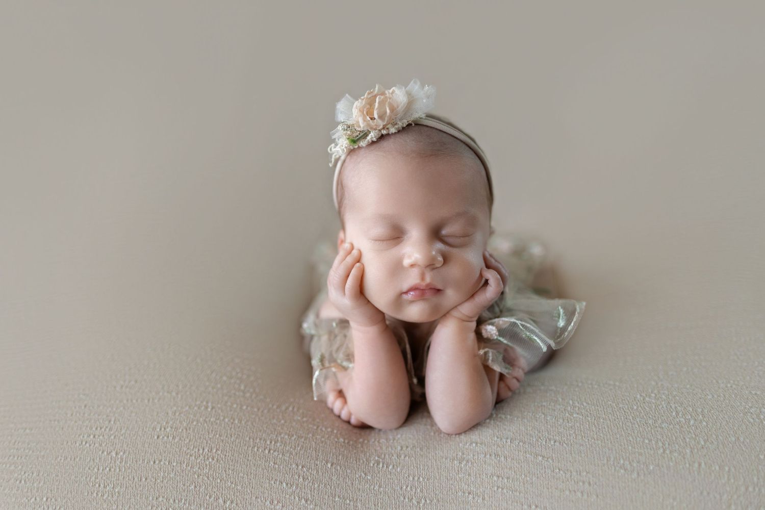 Newborn baby girl sleeping peacefully in froggy pose wearing floral headband and green outfit on beige backdrop.