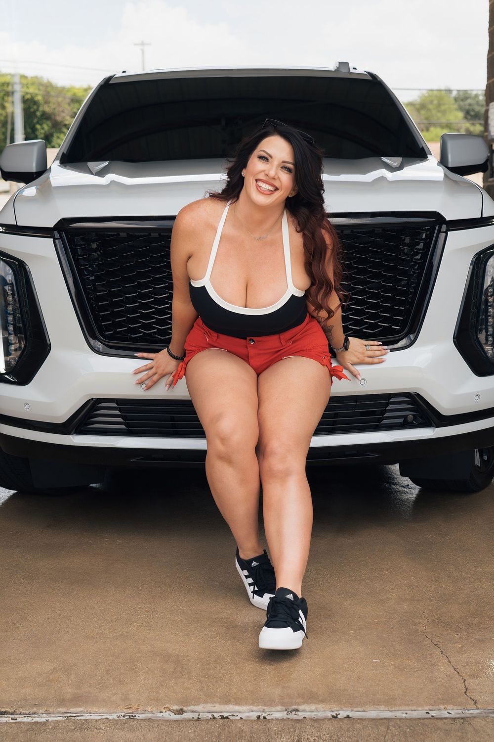 Smiling woman in red shorts and black top posing on white SUV hood in parking area.