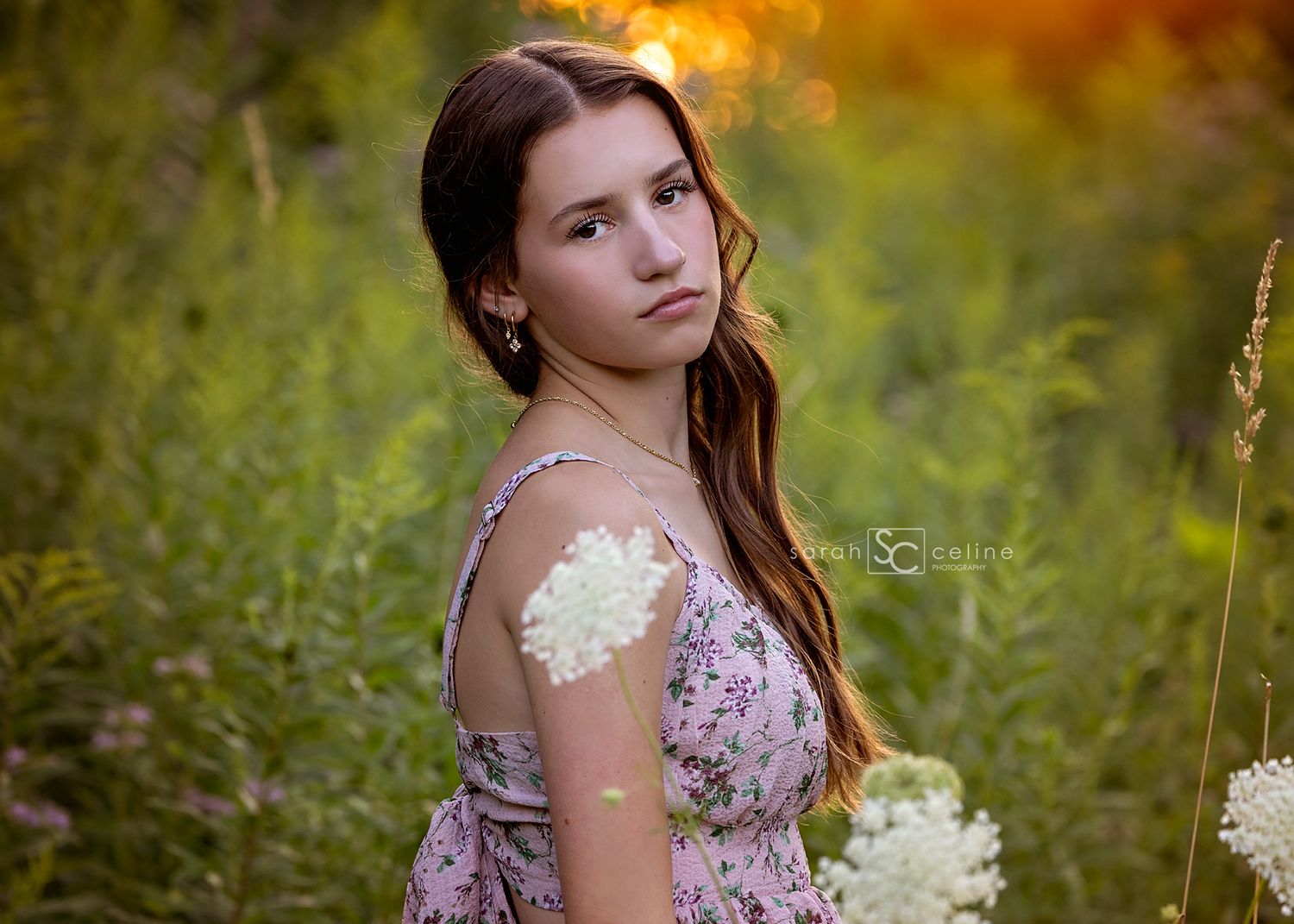 Teen girl in floral dress holding white flowers in a sunlit green field during golden hour photography session.