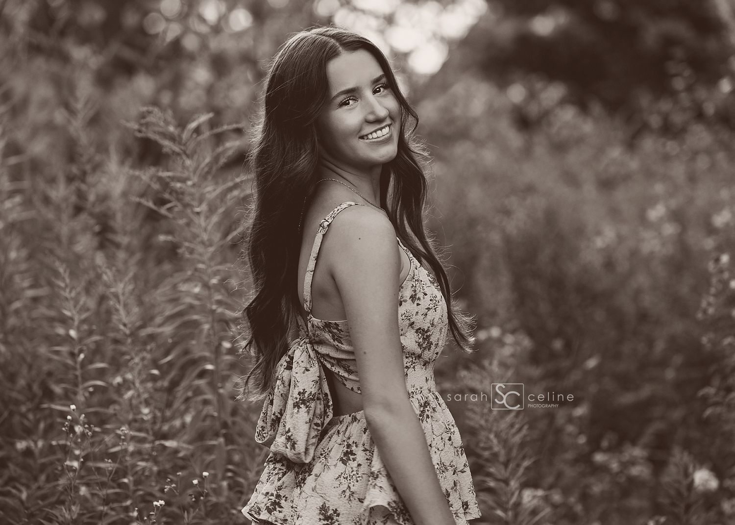 Black and white portrait of a smiling young woman in a floral dress standing among tall wildflowers outdoors.