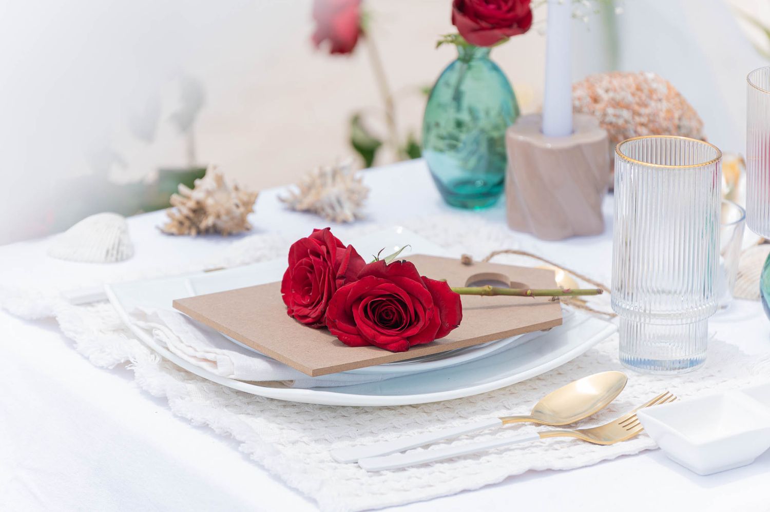 Elegant table setting with red roses on a plate, gold cutlery, and teal vases on a white tablecloth.