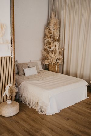 Minimalist boho bedroom with white bedding, pampas grass decor, and warm wood flooring in a neutral color scheme.