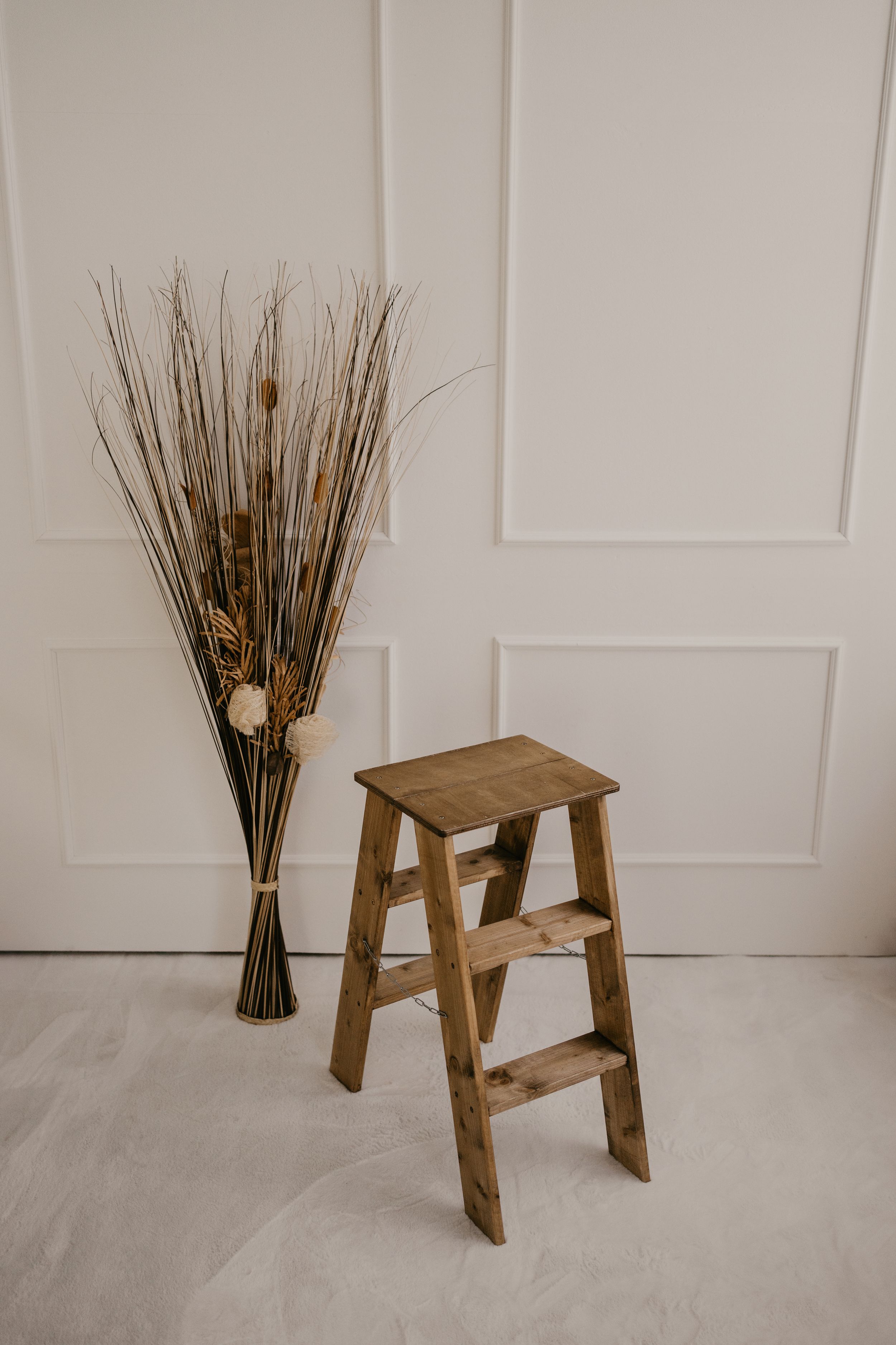 A sequence showing a wooden step stool being placed next to dried floral arrangements against a white wall.