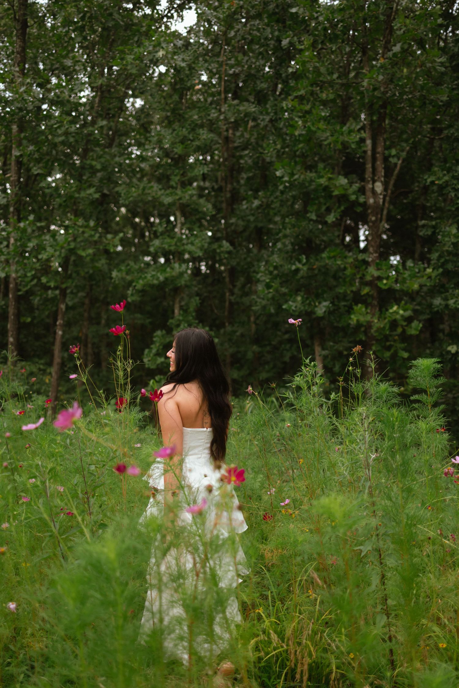 Woman in white dress standing in wildflower meadow surrounded by lush green forest trees.