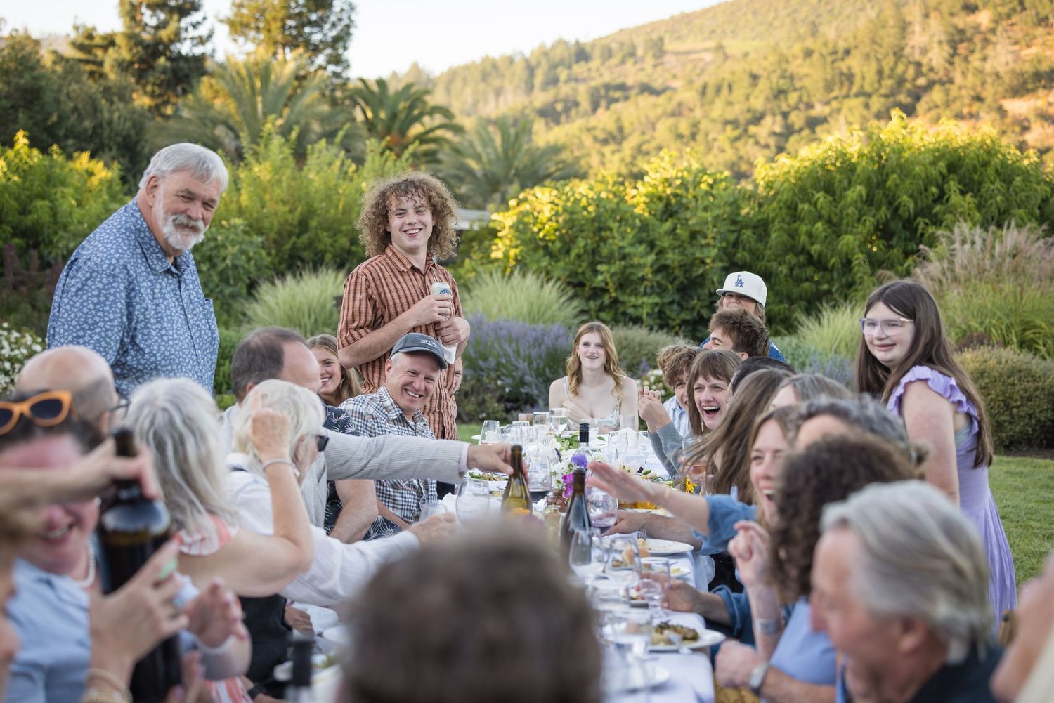 Large group of people enjoying an outdoor dinner party at a long table surrounded by lush greenery at sunset.