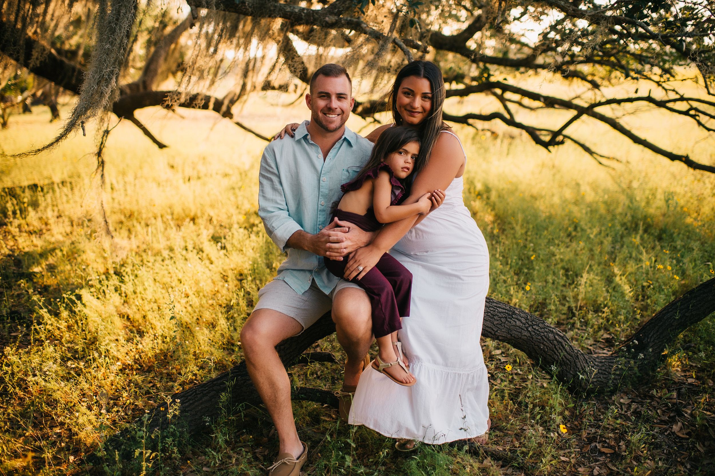 Happy family of three posing outdoors under a tree in a sunlit meadow, parents embracing their young child.