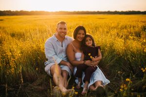 Family of three sitting in golden field at sunset, smiling couple with young child surrounded by yellow wildflowers.