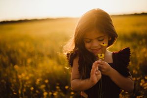 Young girl smelling wildflowers in golden sunset field, wearing dark dress with ruffled sleeves.