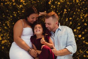 Family of three lying in sunflower field, parents smiling at laughing girl in red dress.